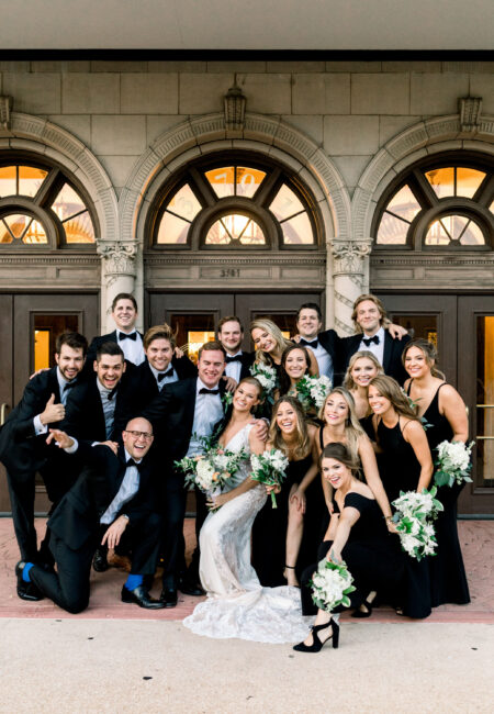 14 person wedding party in front of stone building, men in black tuxes and women in black dresses,