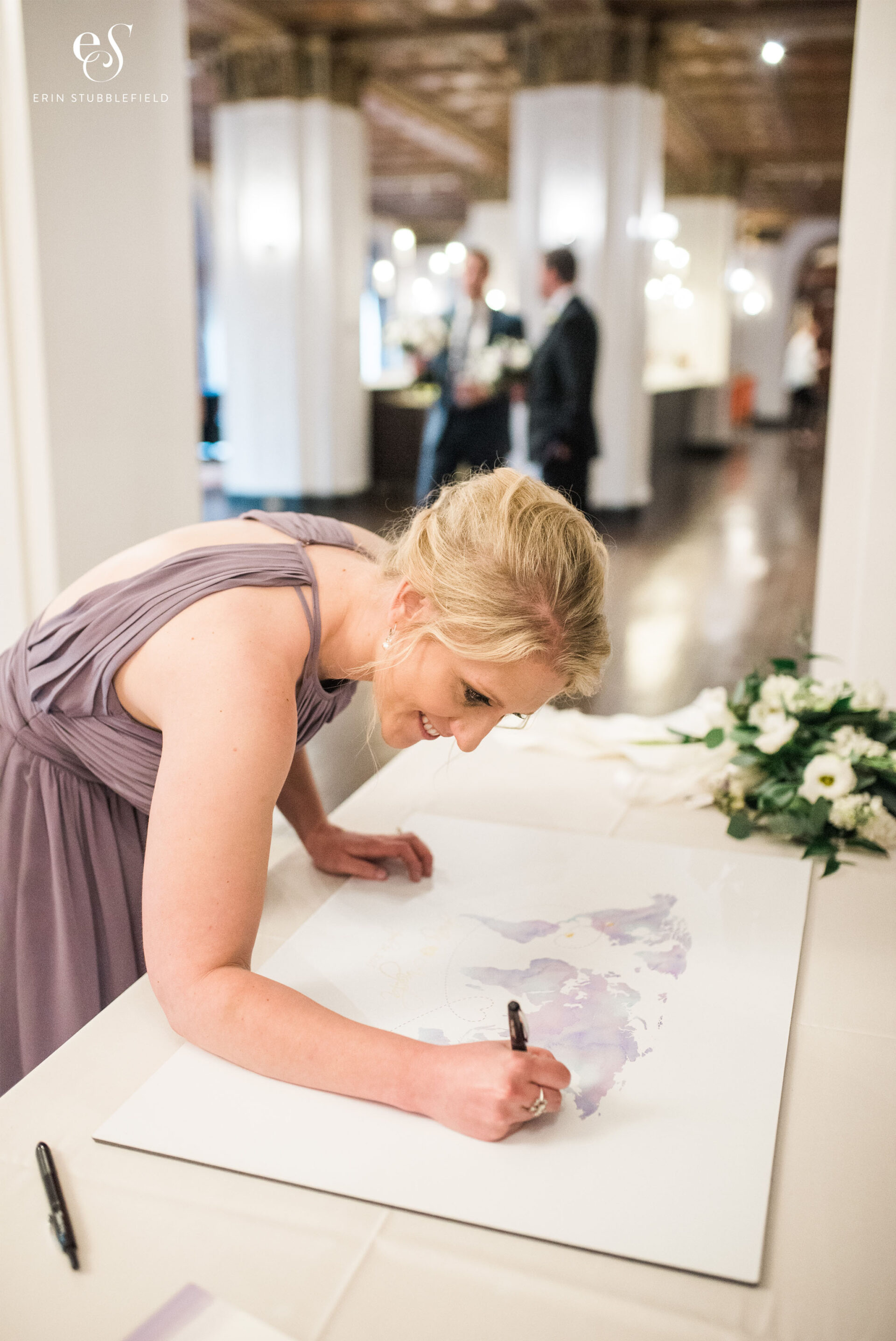 Bridesmaid in a light pink dress signing over her home city in wedding guest book, which is actually a map