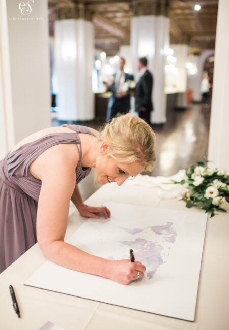 Bridesmaid in a light pink dress signing over her home city in wedding guest book, which is actually a map