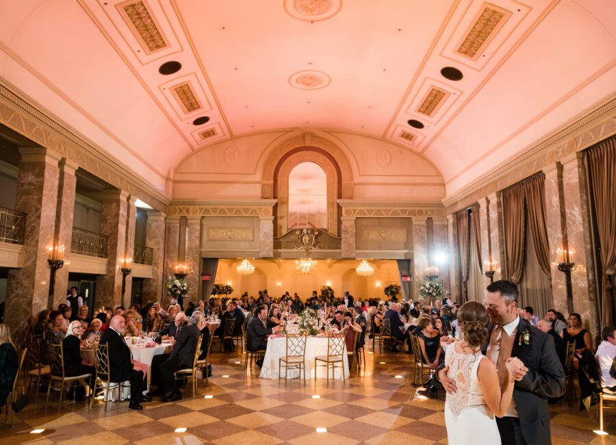 Straight wedding couple's first dance in front of onlooking guests in a pink-lit ballroom