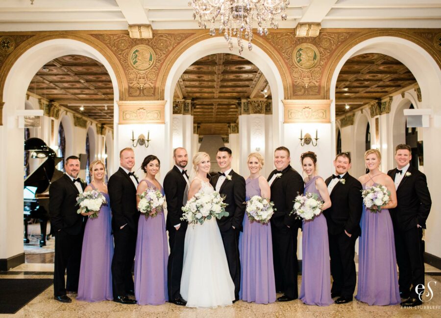 12 person wedding party in elegant room, with suited men and purpled-dressed women rotating and wedding couple in the center