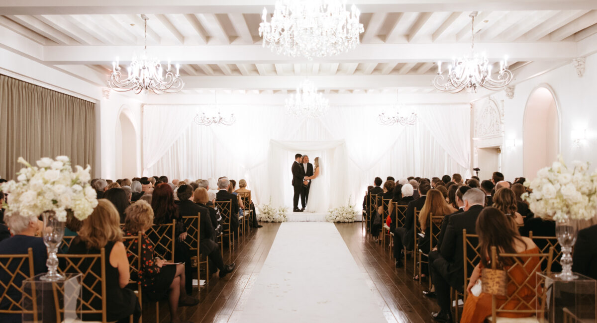 A couple stands at the altar during a wedding ceremony in an elegant, chandelier-lit hall.